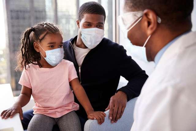 A doctor wearing goggles and mask is facing a young girl and dad with masks.